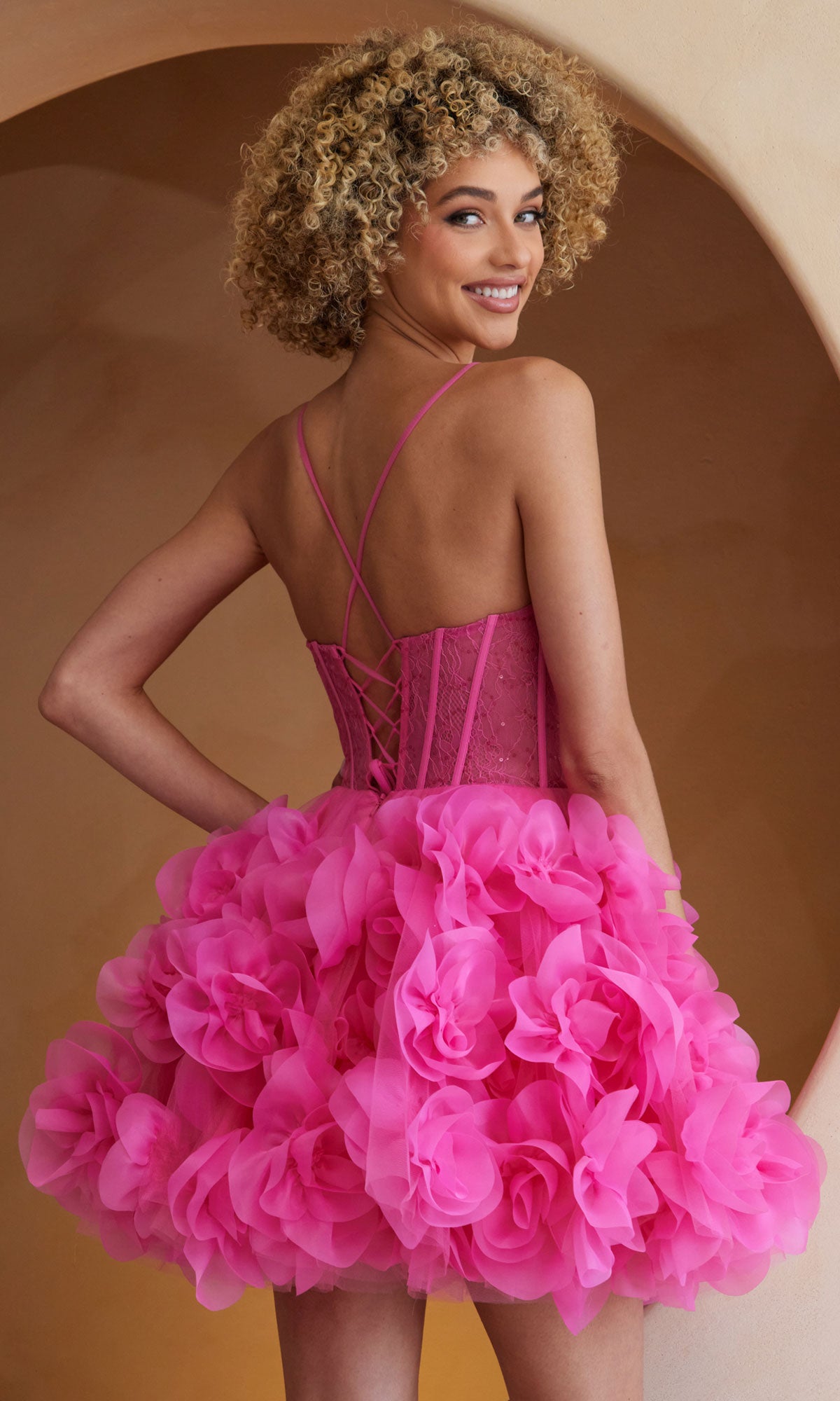 A woman wears the Amelia Couture SU083S short pink homecoming dress with a ruffled flower-like skirt and lace-up back, standing before a curved beige background.